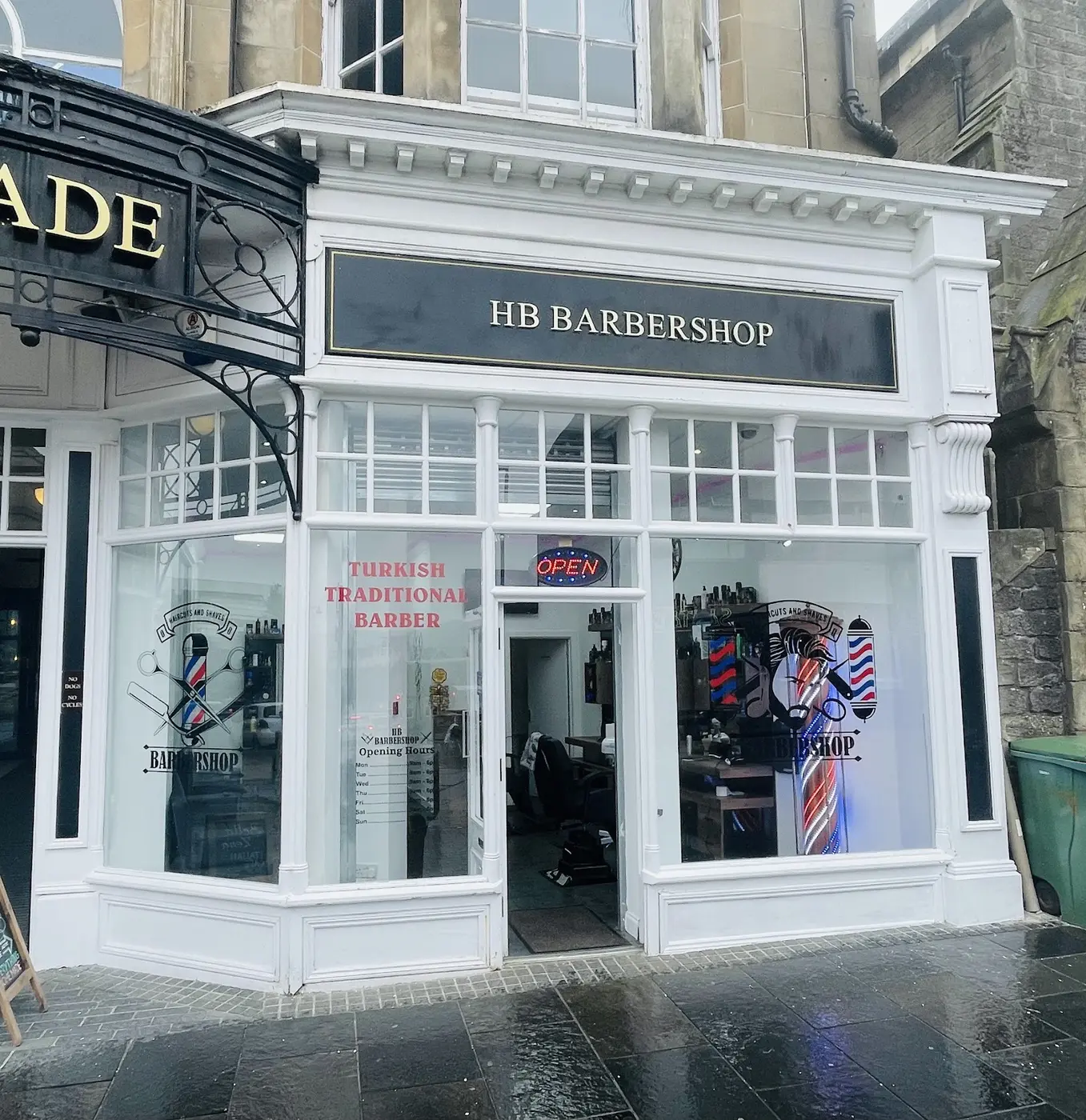 The black and gold HB Barbershop shopfront with white woodwork, an OPEN sign and barber pole graphics in the windows